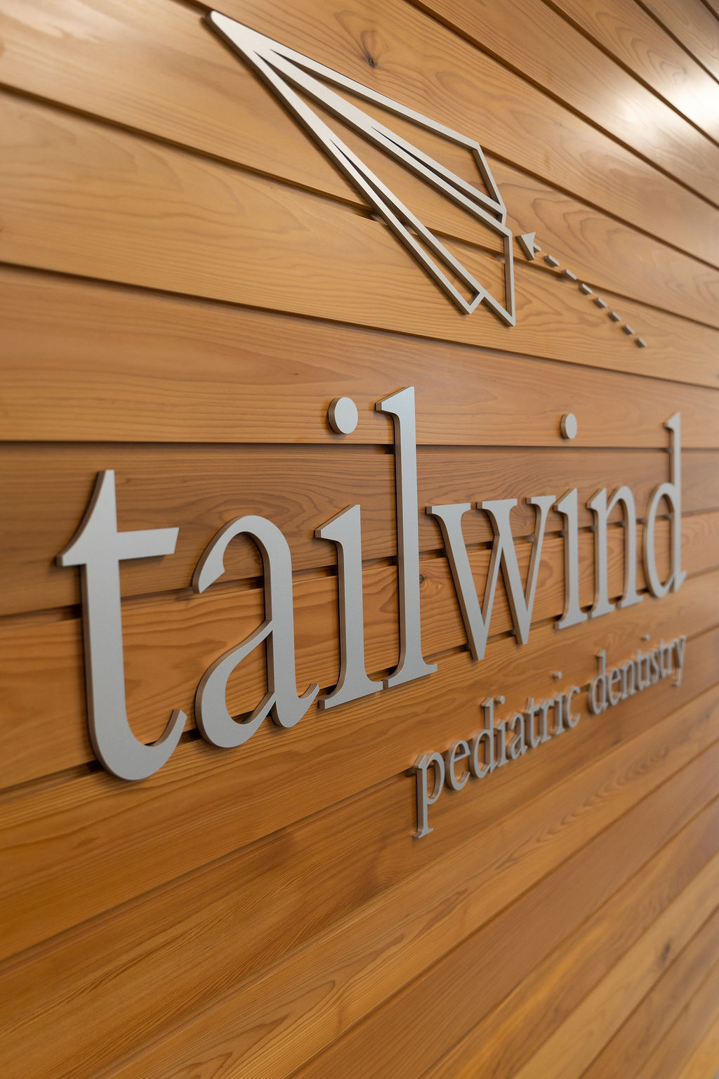 Wooden wall with silver "tailwind pediatric dentistry" sign and paper plane logo, suggesting a child-friendly dental practice with a modern aesthetic.
