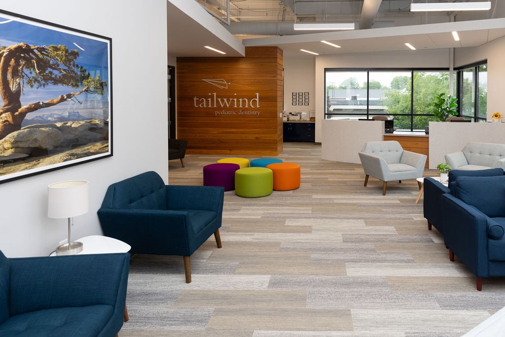 Modern office lobby with blue chairs, colorful round stools, and large windows. "Tailwind Pediatric Dentistry" sign on wood-panel wall. Bright, inviting space.