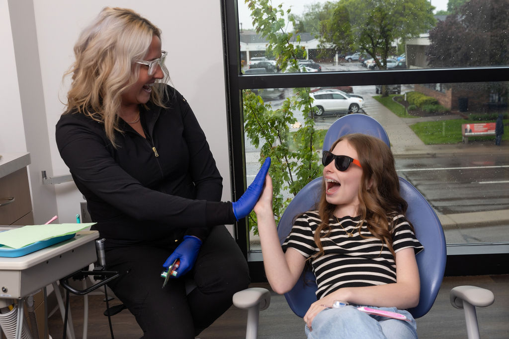 A person in black attire gives a high-five to a smiling young individual in sunglasses, seated in a dentist chair by a window.