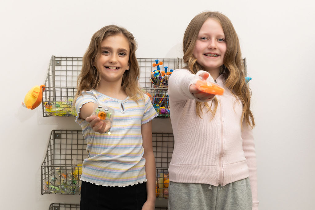 Two children smiling and holding toys stand in front of a wall-mounted rack filled with colorful small toys against a plain background.