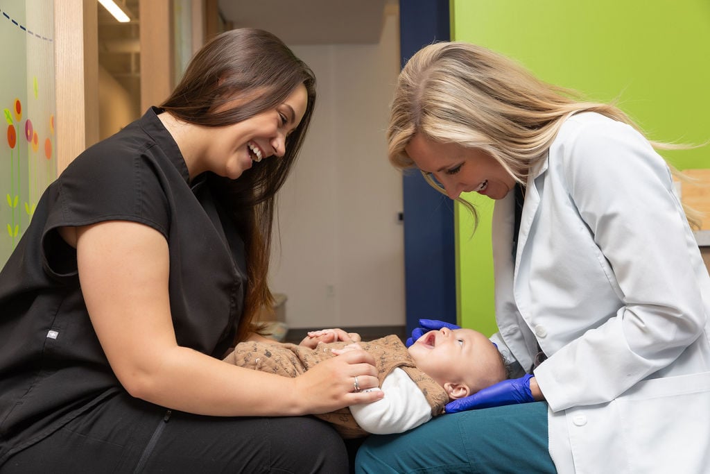 Two people smiling at a baby lying on an examination table in a bright, cheerful room with green walls and colorful decorations.