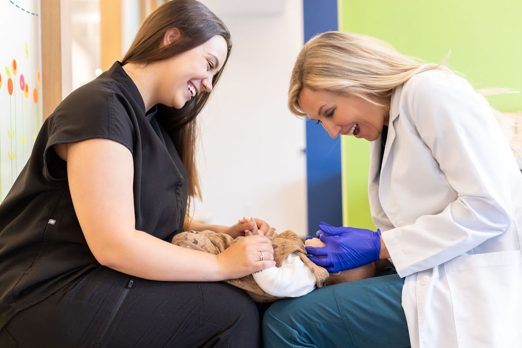 Two persons, one in a lab coat and gloves, smiling at a small animal in a veterinary clinic setting.
