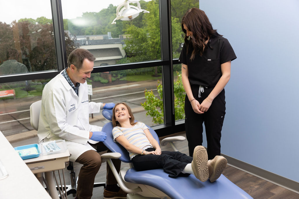 A person in a white coat and another in scrubs attend to a smiling child in a dental office with large windows.