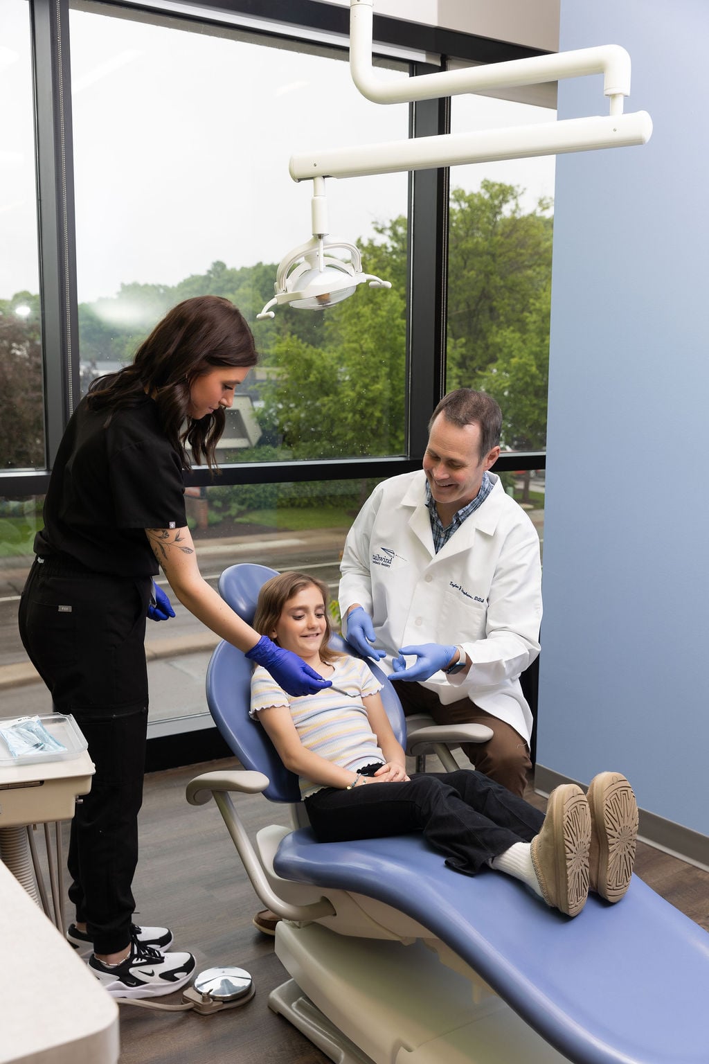 Two persons in dental uniforms attend to a smiling child patient in a dentist chair. Large windows reveal a green, tree-filled view.