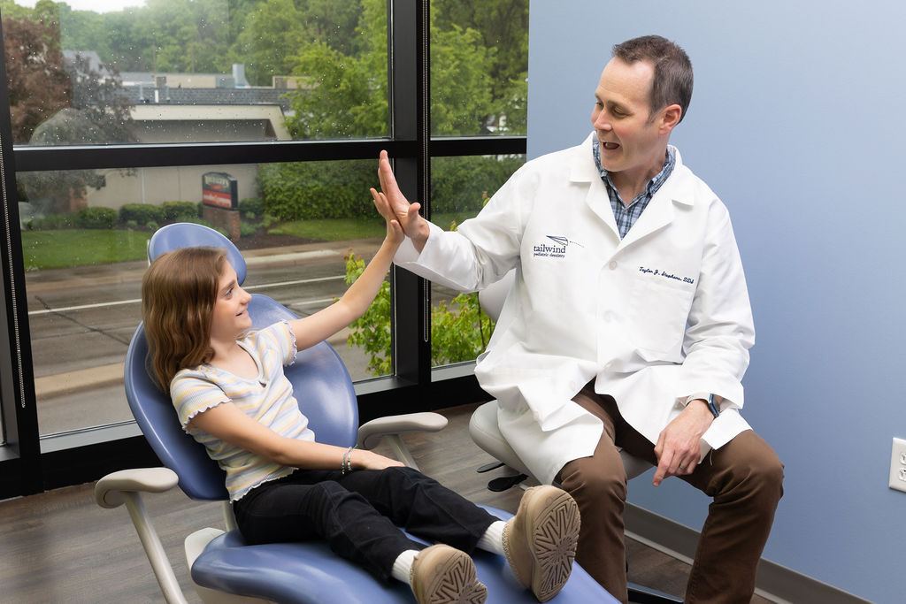 A person in a white coat and a child share a high-five in a dental office. Large window shows greenery outside.