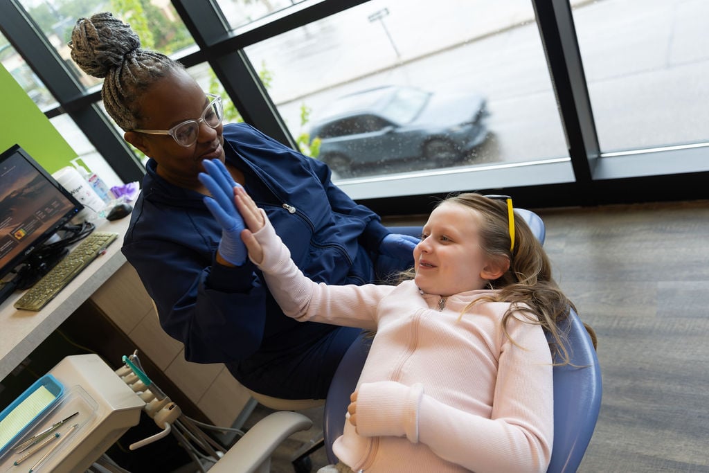 A person in a dental office high-fives a child seated in a dental chair, with equipment and large windows in the background.