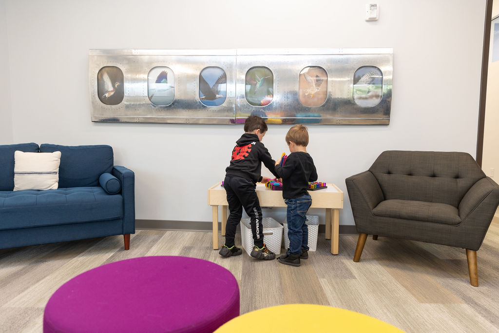Two children play with blocks in a modern waiting area, featuring colorful furniture and an airplane window-themed wall art.