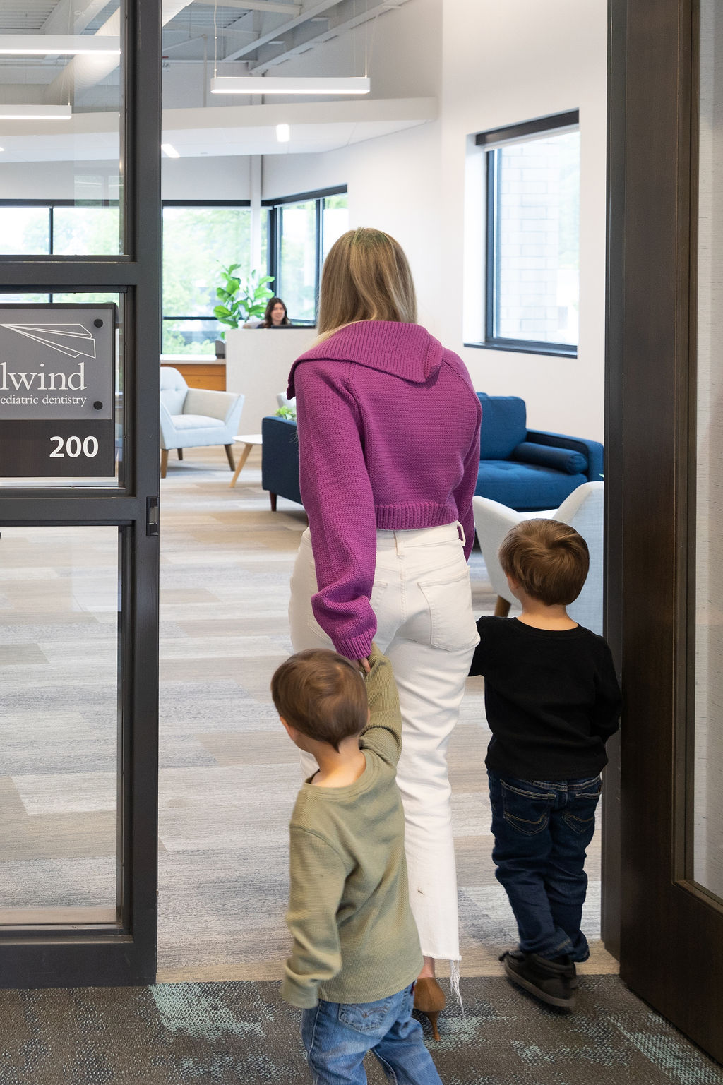 A person with two children enters a modern dental office, featuring large windows and comfortable seating in a welcoming lobby area.
