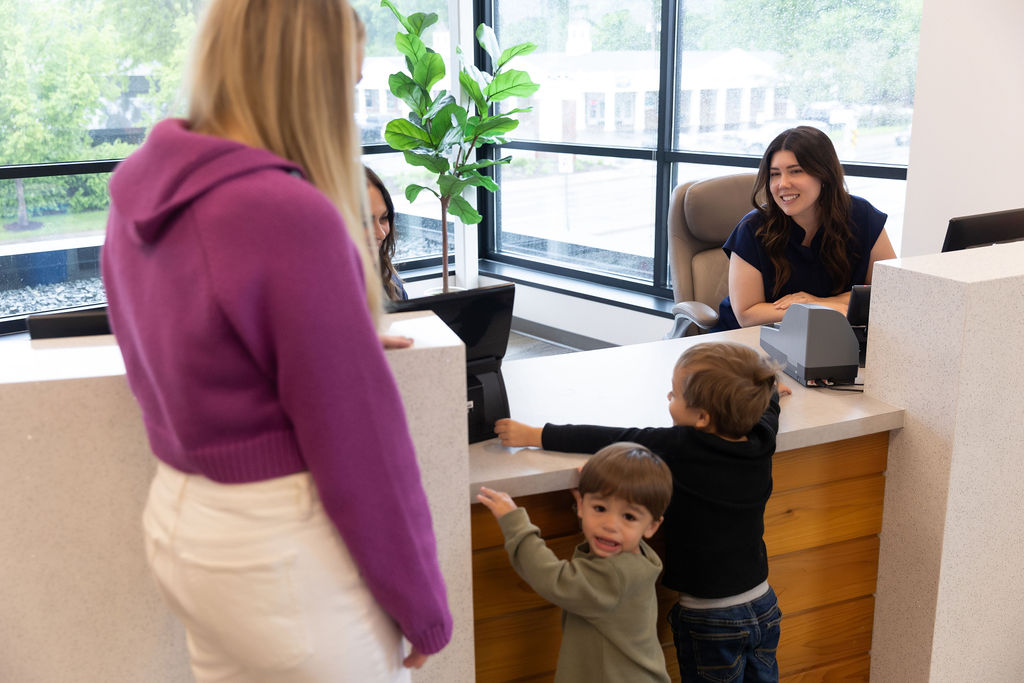 Two children and a person engage with a receptionist at a counter. Large windows and a potted plant decorate the modern space.