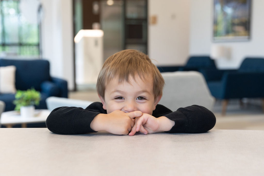 A child smiles playfully over a table in a cozy, modern living room with blue sofas and natural light streaming in.
