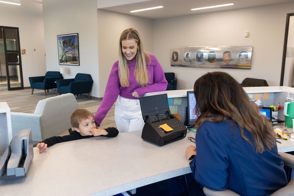 A person and child at a reception desk interact with another person in an office, with chairs and art in the background.