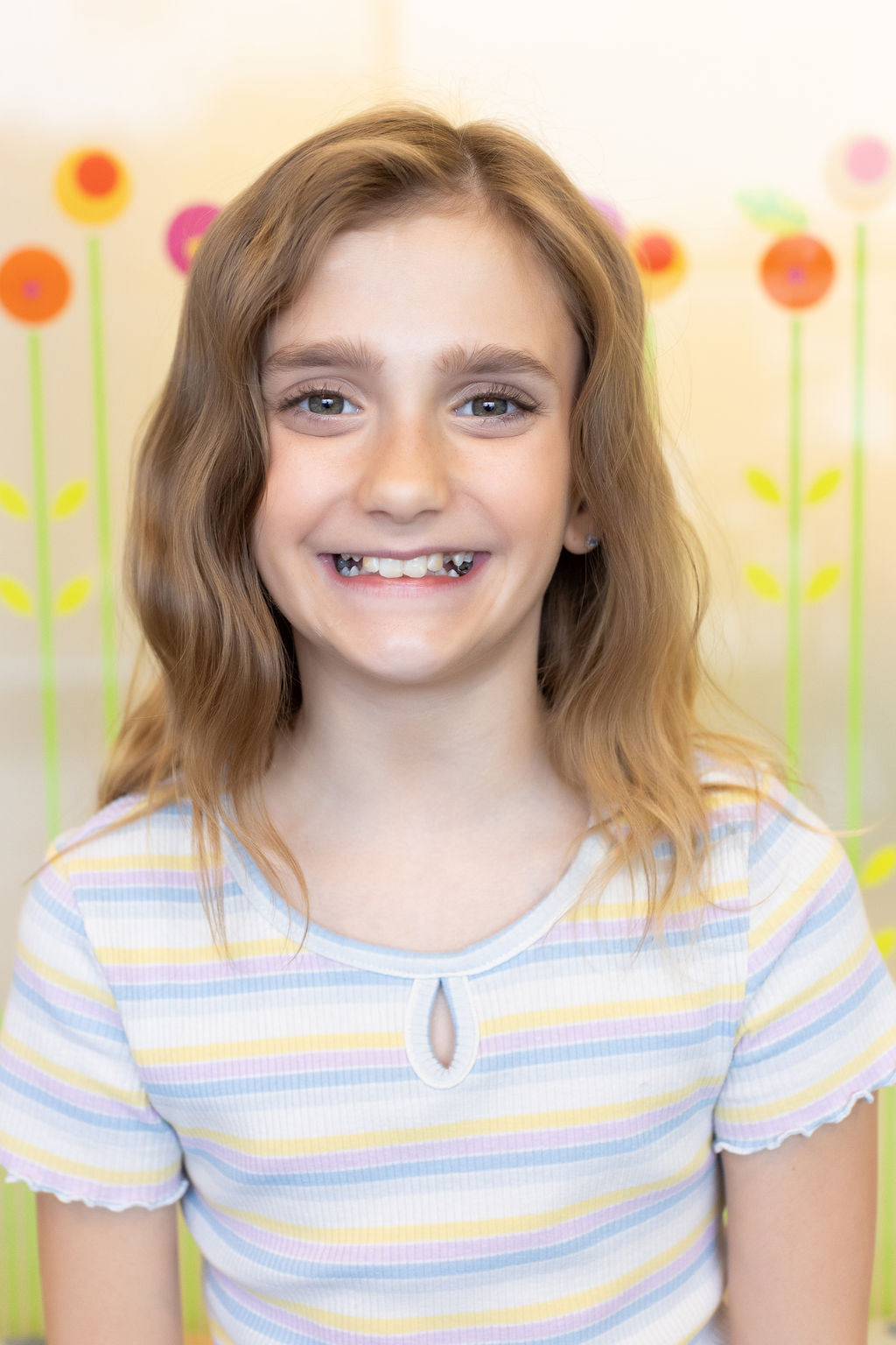 A smiling child with long hair is in front of a colorful floral mural, wearing a striped shirt indoors.