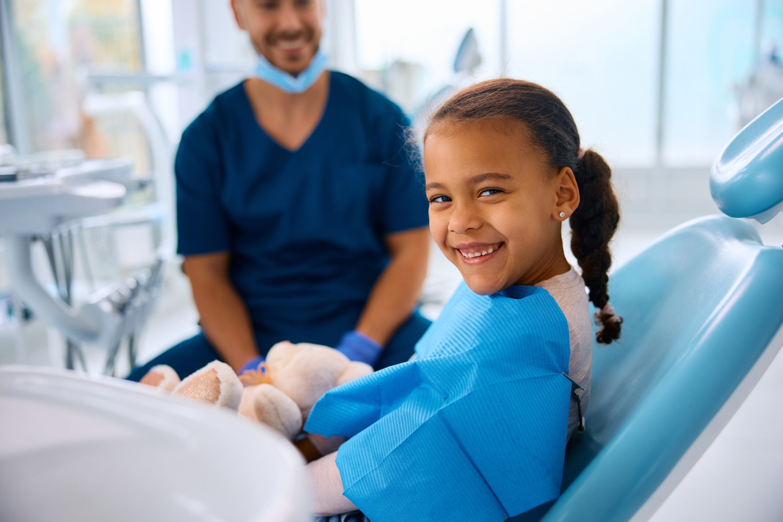 A smiling child holds a stuffed bear in a dental chair, with a person in scrubs nearby in a bright, modern dental office.